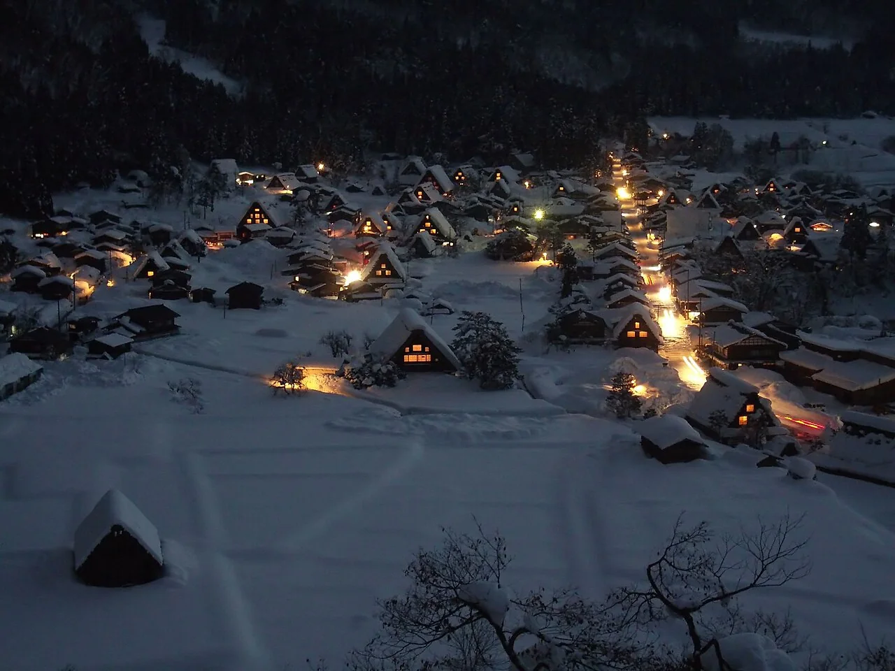 Casas de Shirakawa-go iluminadas à noite entre árvores e telhados cobertos de neve.